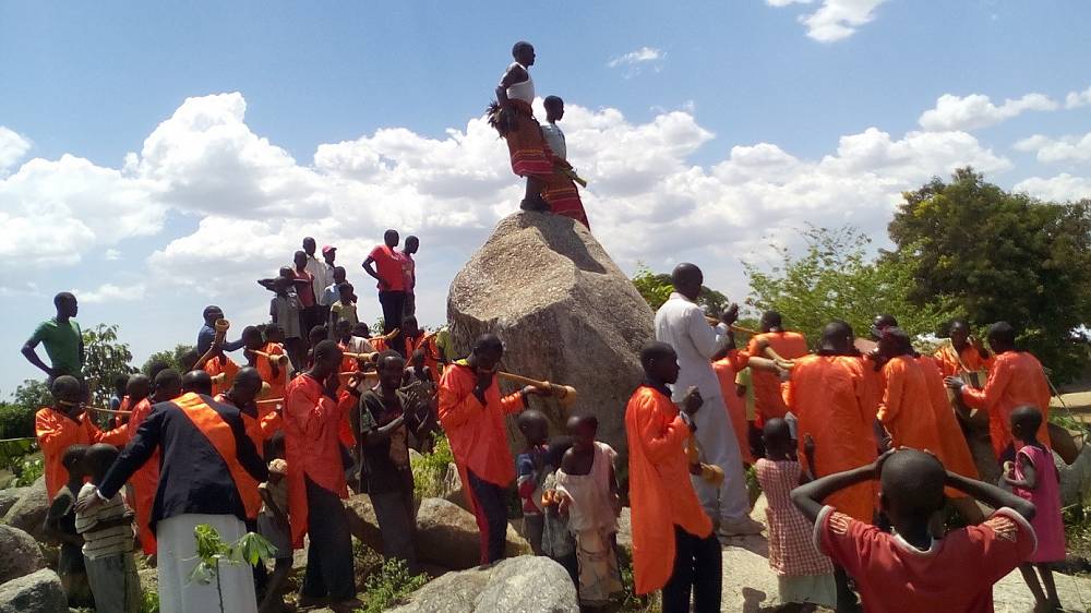 Uganda Culture Guide 15 Basoga people in traditional orange attire gathering around a large rock.