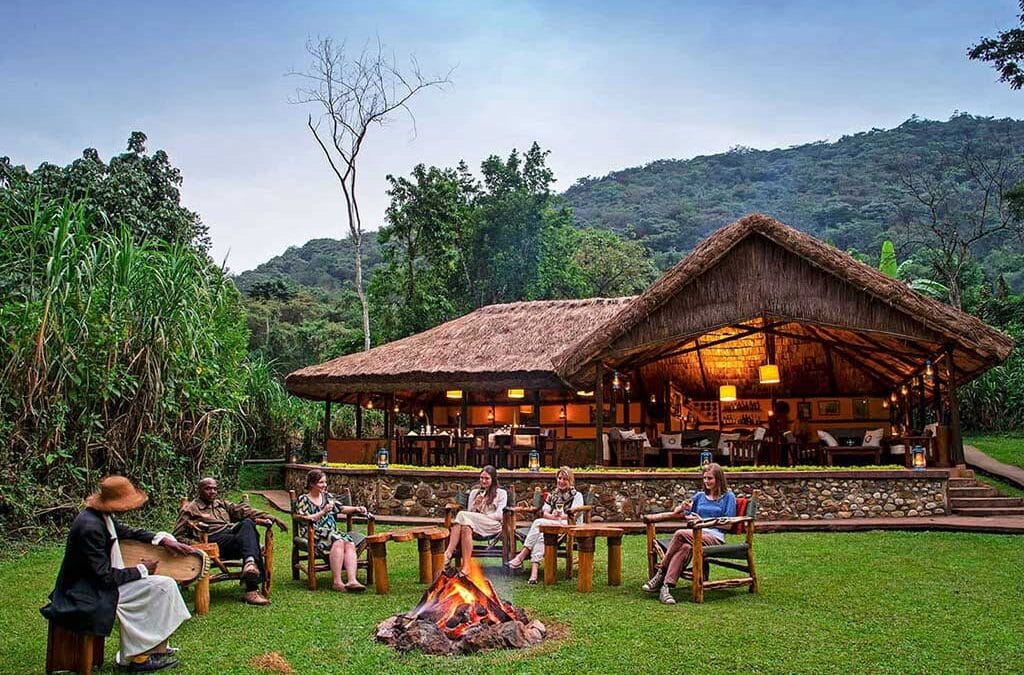 Luxury Gorilla Lodges in Bwindi 7 Tourists relaxing in safari chairs around an outdoor campfire on a green lawn in front of a thatched-roof luxury lodge in Bwindi