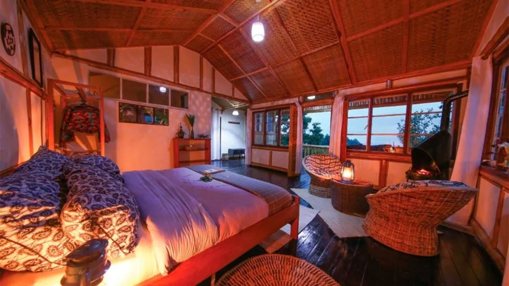 Luxury Gorilla Lodges in Bwindi 19 The warmly lit interior of a luxury safari cabin bedroom featuring a large bed with blue and white patterned linens wicker chairs and a small fireplace under a woven mat ceiling at dusk