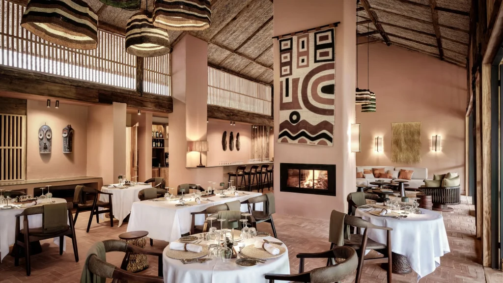 Luxury Gorilla Lodges in Bwindi 17 An elegant upscale safari lodge dining room with pink-toned clay walls a double-sided fireplace and beautifully set tables with crisp white linens