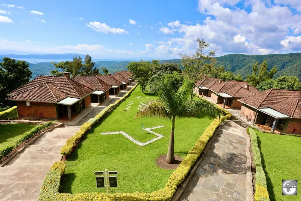 Nyungwe Chimpanzees 45 A high-angle view of two symmetric rows of brick cottages with terracotta tile roofs facing a manicured green lawn with a palm tree, set against a backdrop of rolling green hills and a blue sky.